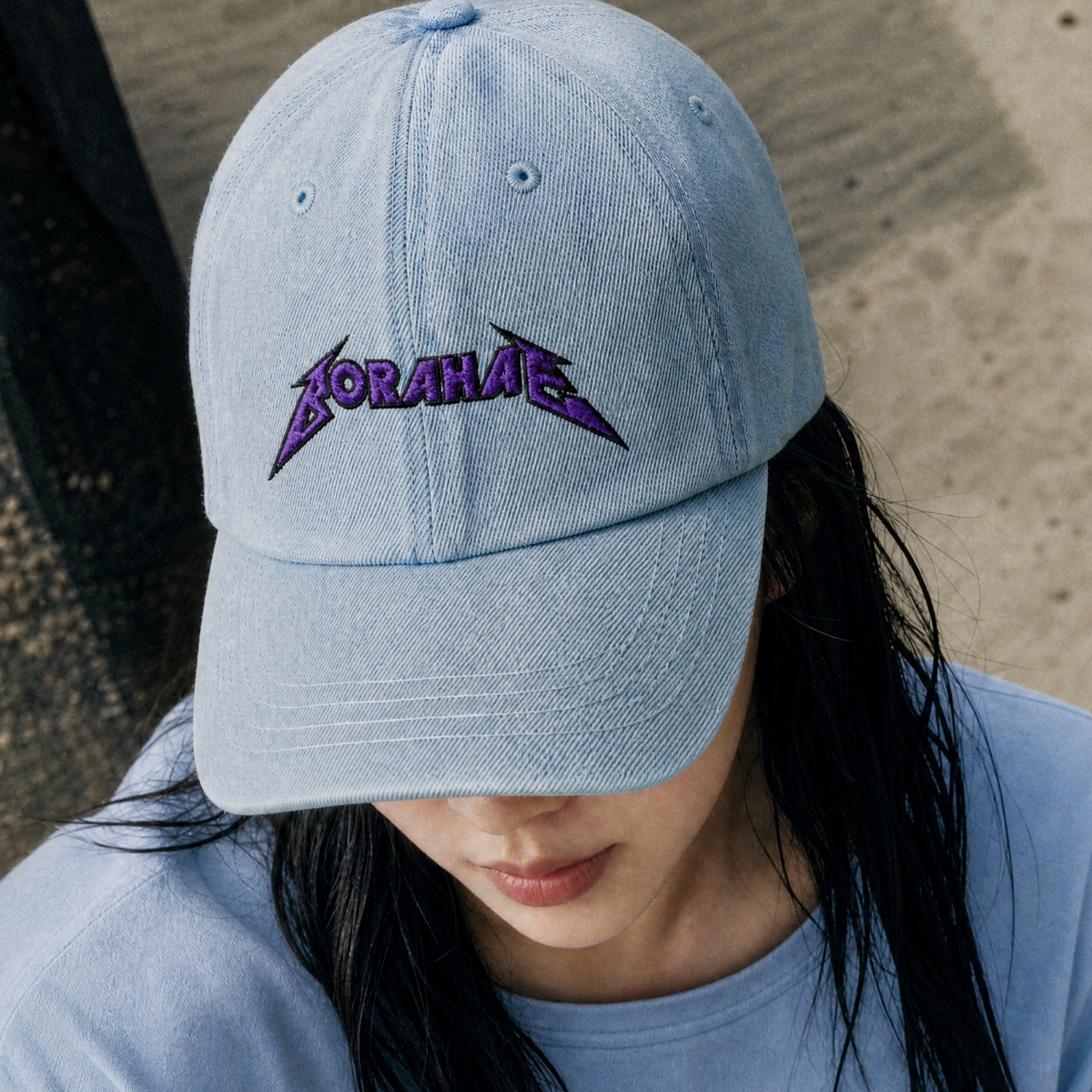 A person wearing a light blue baseball cap with purple embroidered text reading 'BORAHAE' across the front, photographed outdoors against a blurred concrete background.